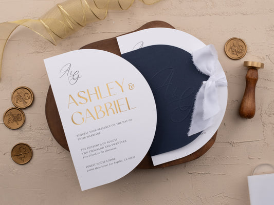 Navy blue wedding invitation with gold foil print and white ribbon, arch-shaped layout styled on neutral background