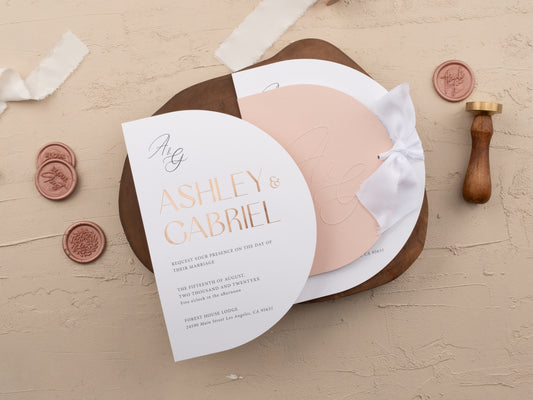Blush pink wedding invitation with rose gold foil print, half-moon design and white ribbon, shown on a neutral background