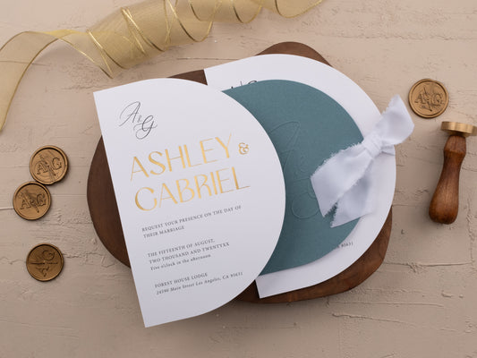 Dusty blue wedding invitation with gold foil print, arch-shaped design and white ribbon, styled on neutral background